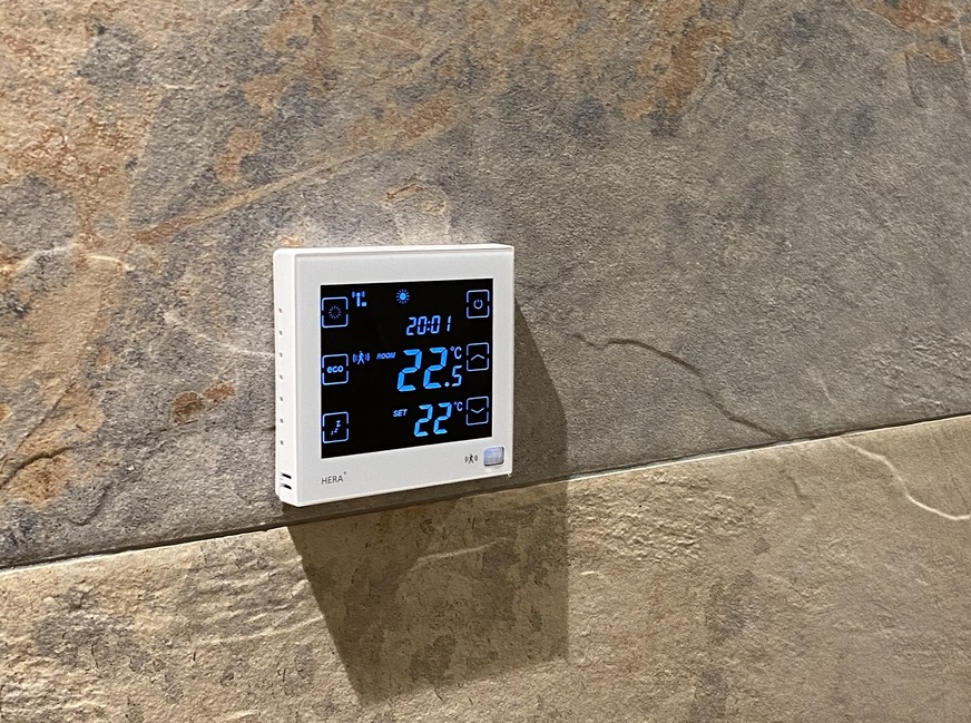 Blossom-ic Funk-Raumthermostat Hera Direct+ weiss (230V) mit Anzeige der Uhrzeit, Soll- und Ist-Temperatur und dem aktuellen Heizprogramm. Das Hera+-System kann manuell über die Funk-Raumthermostate und/oder über die Blossomic-App geregelt werden.