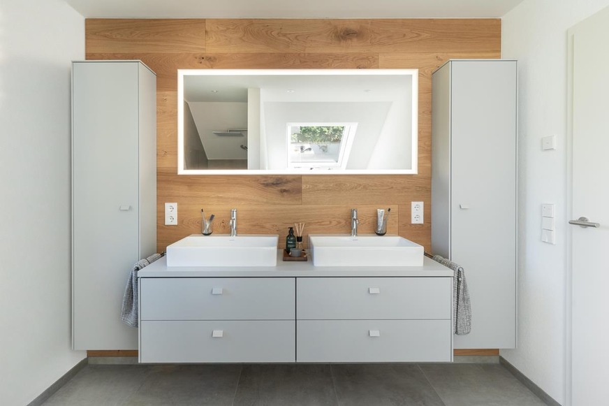 Möbel in hellem Grau, zwei Waschbecken, ein großflächiger Spiegel und als Highlight die Waschtischrückwand aus massiver Eiche.