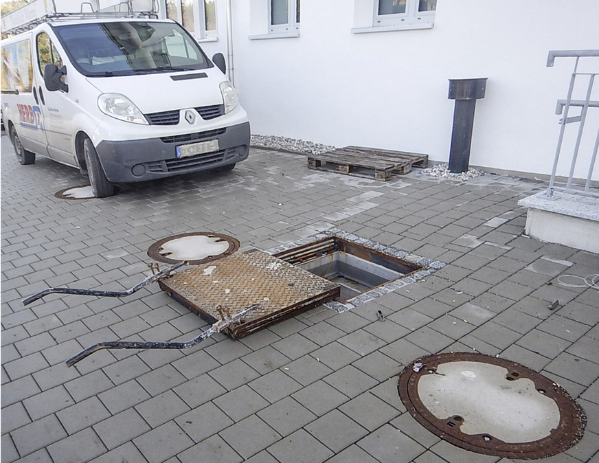 Blick auf die drei Befüllöffnungen und auf den rechteckigen Behälter­einstieg nach der Fertigstellung der befahrbaren Fläche über dem Pelletspeicher. An der Hauswand sichtbar die mit dem Speicher-Innenraum verbundene Lüftungsleitung DN 200, die ca. einen Meter über Gelände mit einer schlagregensicheren Haube abgedeckt ist.