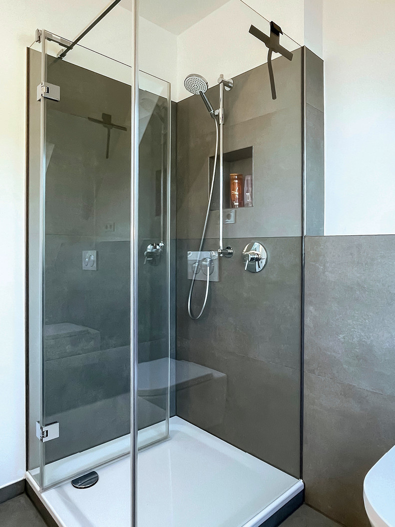 Die Duschabtrennung besteht aus einem Festglas-Element an der Seite und vorne einer Tür mit Pendeltürbeschlag neben einem kleinen Festteil.
