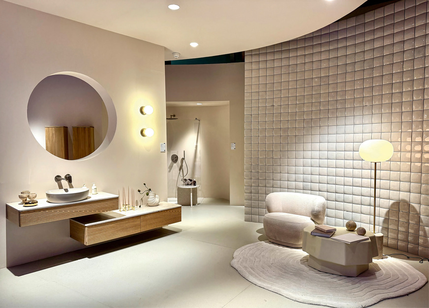 Statt durch Farbe fällt das Bad durch geometrisch gerasterte Gestaltung des Reliefs auf der geschwungenen Wand auf. Warmes Off-White, Teddyfell auf dem Polster und helles Holz machen die unaufdringliche Einrichtung gemütlich. ­Gesehen auf der ISH 2025 bei Villeroy & Boch.