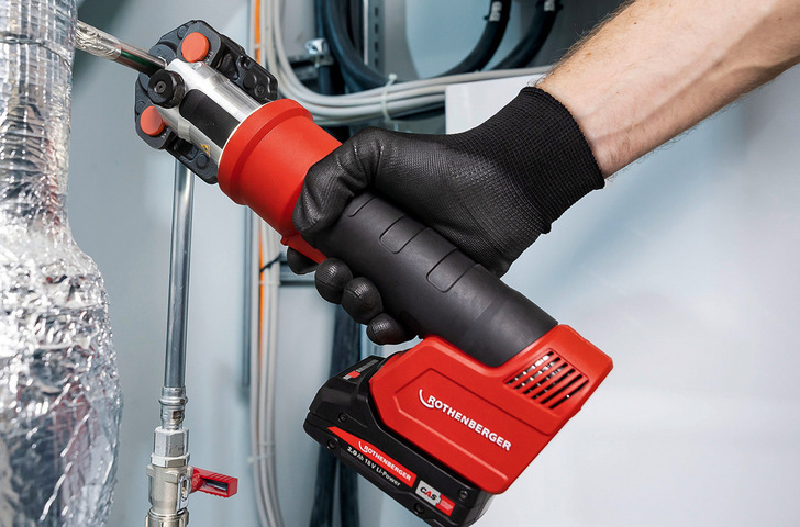 Gerne! Hier der optimierte, themenrelevante Alt-Text für das beschriebene Bild: Alt-Text: „Eine Handwerkerin oder ein Handwerker mit Schutzhandschuhen setzt in einer Industrieumgebung ein rotes Rothenberger Presswerkzeug an, um eine Metallrohrverbindung fachgerecht zu verpressen – typischer Arbeitsablauf bei der Installation von Heizungs-, Sanitär- und Trinkwasserleitungen im SHK-Bereich. - © Bild: Rothenberger