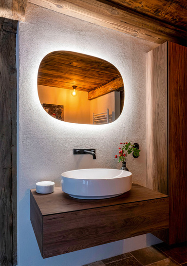 Der ovale „Geberit Option Lichtspiegel“ bietet ein angenehmes Licht. Der Vari-Form-Aufsatzwaschtisch und die „One Wandarmatur“ in Schwarz matt fügen sich in die Gestaltung des Bades ein.