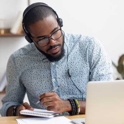 Focused african business man in headphones writing notes in notebook watching webinar video course, serious black male student looking at laptop listening lecture study online on computer e learning