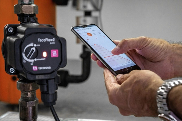 Bildbeschreibung: Ein Installateur nutzt eine Smartphone-App zur Steuerung einer energieeffizienten TacoFlow2 Pumpe, die neben modernen Rohrleitungen in einem Heizungskeller installiert ist. - © Taconova