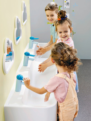 Alt-Text: „Drei Kinder waschen ihre Hände an einem barrierefreien Waschbecken mit höhenverstellbaren, blauen Wasserhähnen, unter wolkenförmigen Spiegeln an einer gelben Wand, symbolisiert die Bedeutung kindgerechter Sanitärlösungen in öffentlichen Einrichtungen.“. - © Bild: Geberit