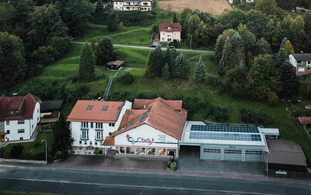 Vogelperspektive eines modernen Wohngebäudes mit rotem Dach, umgeben von üppiger Vegetation. Auf dem Garagendach sind Solarpanel-Installationen zu sehen, die nachhaltige Energielösungen für Heizung und Klimatisierung betonen. - © Bild: Martin Morgenweck