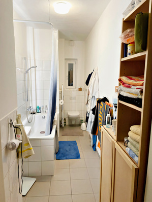 Alt-Text: „Modernes Badezimmer mit platzsparender Installation: Wandhängendes WC neben einer kombinierten Dusch-Badewanne, ergänzt durch hölzerne Regale, die Handtücher und Hygieneprodukte präsentieren – beispielhafte Nutzung effizienter Sanitärtechnik für kleine Räume. - © Bild: Vereinigung Deutsche Sanitärwirtschaft und forsa