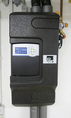 Schwarze Steuereinheit mit digitaler Anzeige und PAW-Logo, montiert an einer weißen Wand, mit oben und unten angeschlossenen Leitungen. - © Bild: Paw