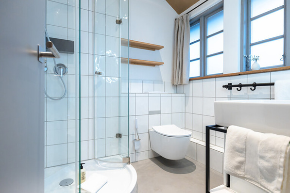Modernes Badezimmer mit Glasdusche, wandmontierter Toilette, Waschbecken, Handtuch und großen Fenstern mit beigen Vorhängen. - © Lüneburger Heide GmbH